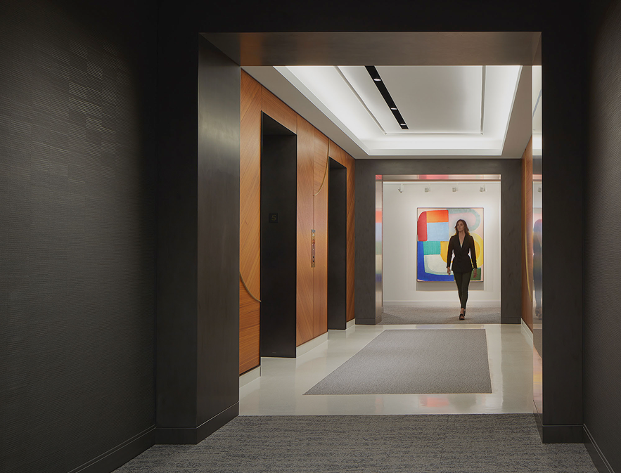 A modern office hallway with wood-paneling and elevator doors, a woman walking toward the camera, and a colorful abstract painting displayed at the far end.